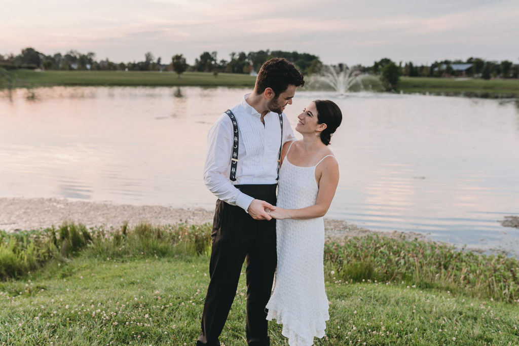 groom smiles at bride at sunset in front of pond with fountain at their charming coxhall gardens wedding