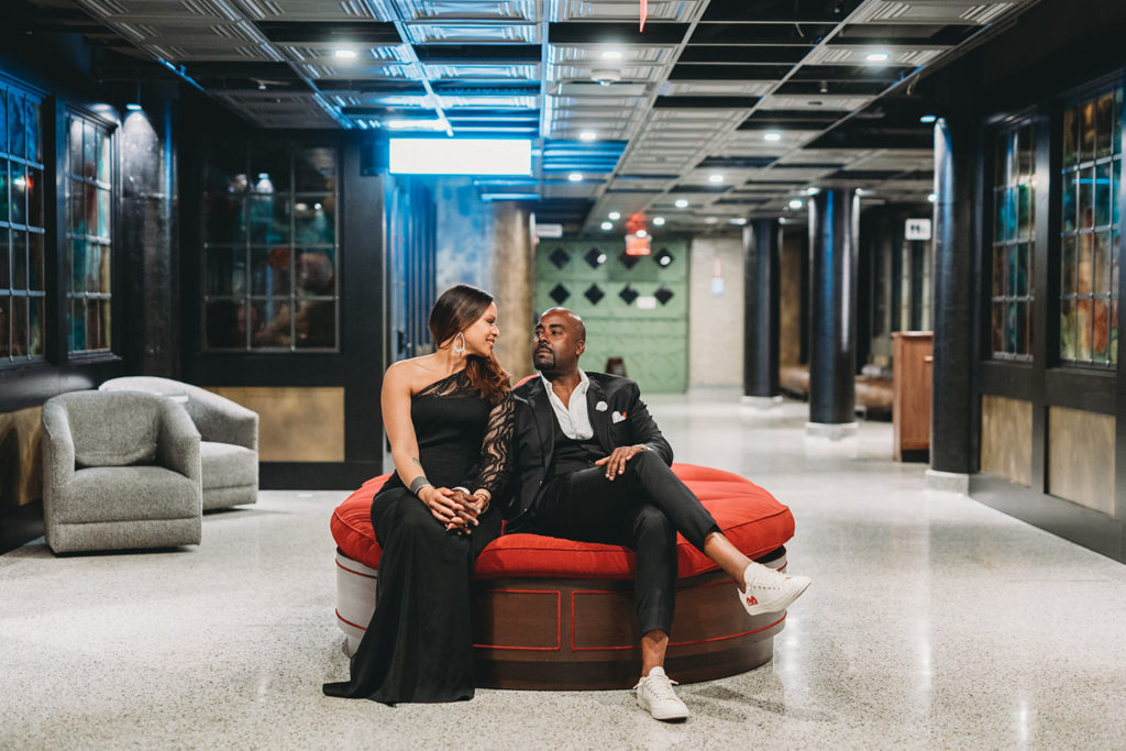 man with crossed legs looks up at woman in ballroom dress from red couch during their photo session