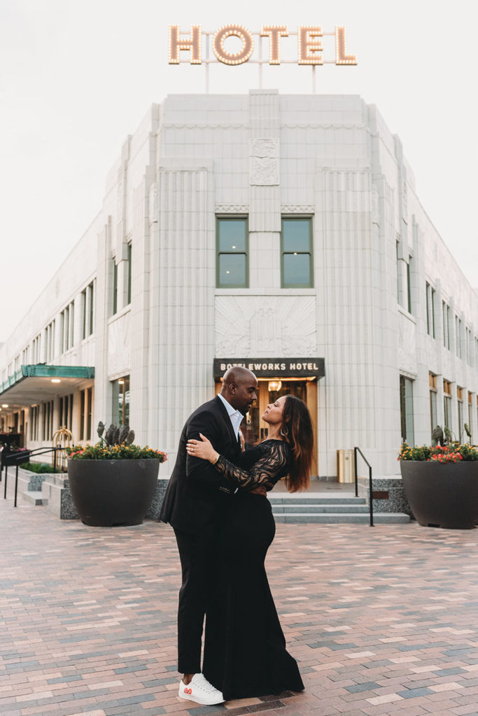 man and woman hug and kiss in front of bottleworks hotel with large hotel sign over the top of them during their photo session