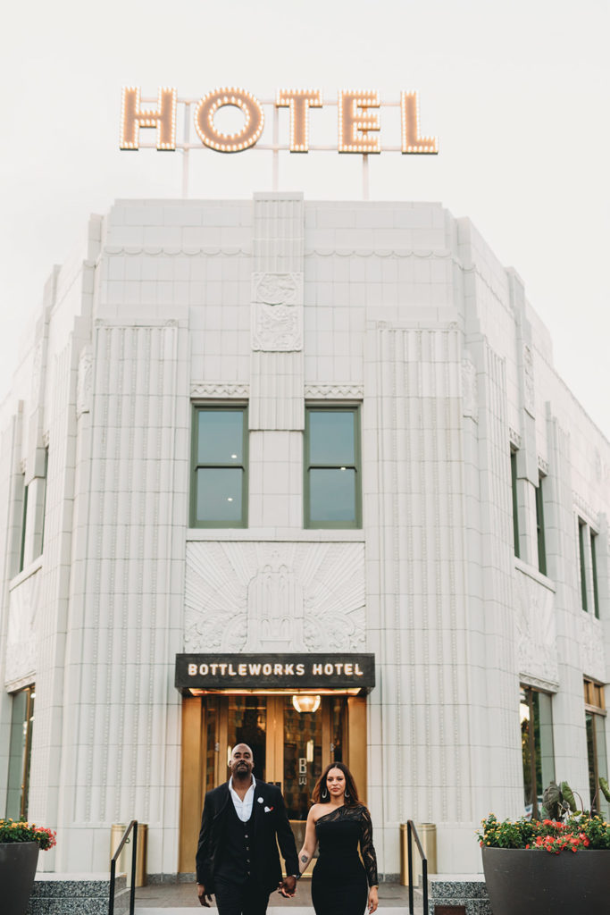 vertical photo of man and woman walking while holding hands in front of the bottleworks hotel under the large hotel sign during their photo session