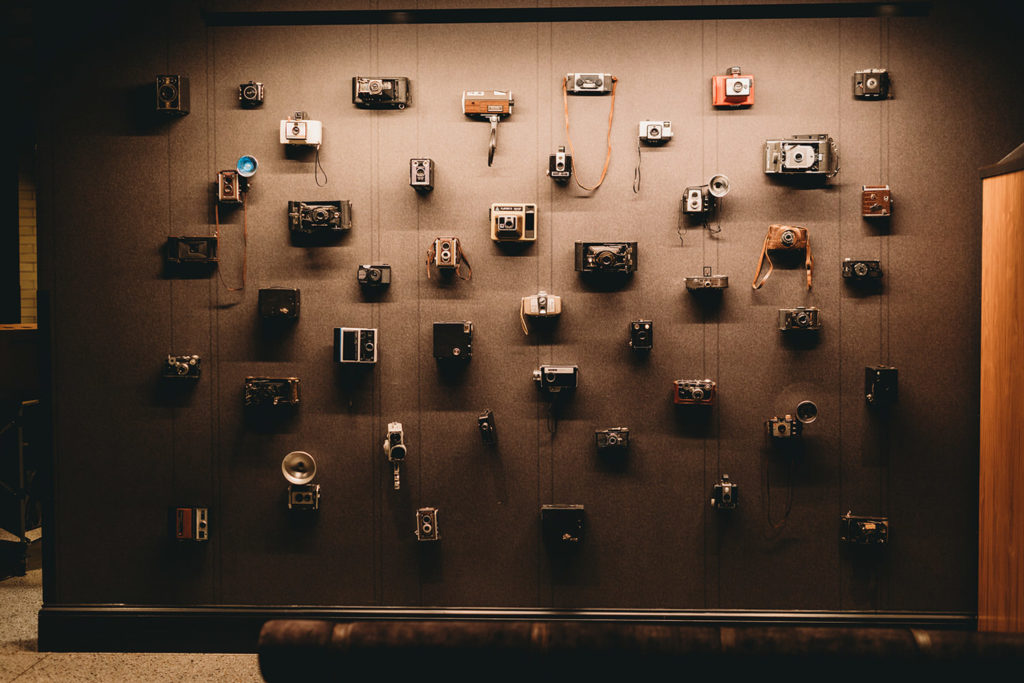 A lot of antique cameras mounted on a wall under a spotlight at the Bottleworks Hotel