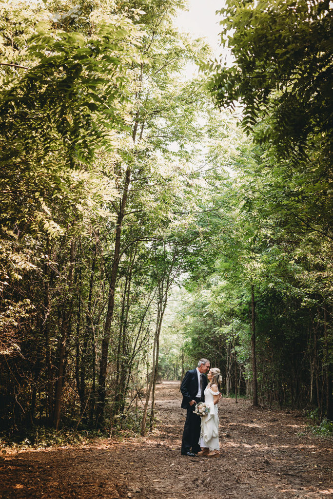 wide shot of groom kissing bride on forehead on wooded path during their five willows wedding