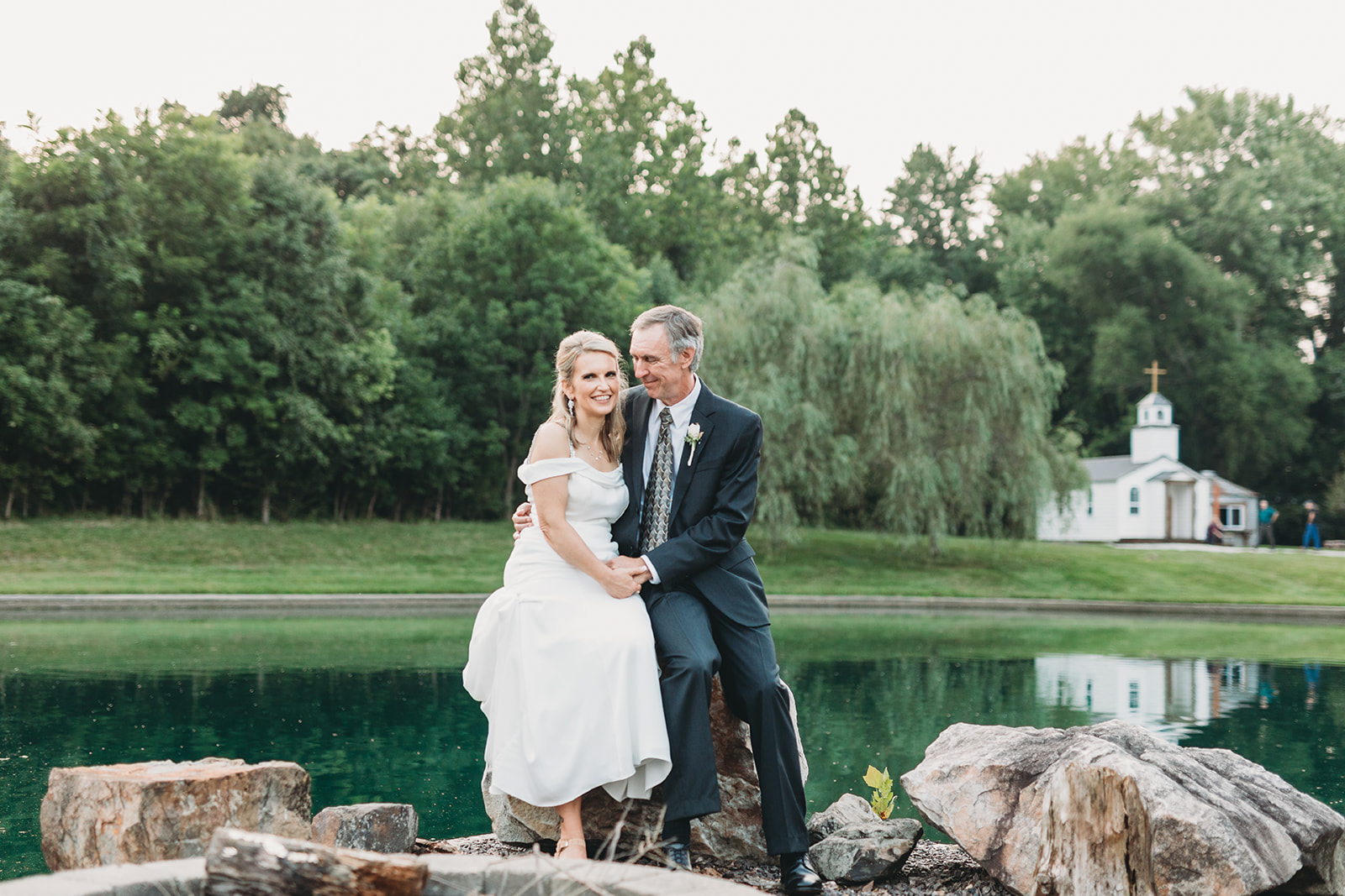 bride and groom sit on rocks in front off pond with small chapel in background during their five willows wedding