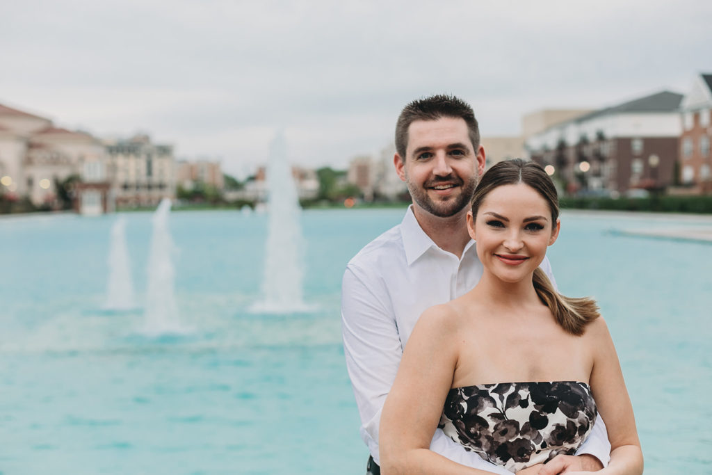 a bearded man hugs a woman in a strapless dress in front of a giant fountain during their Carmel City Center engagement session