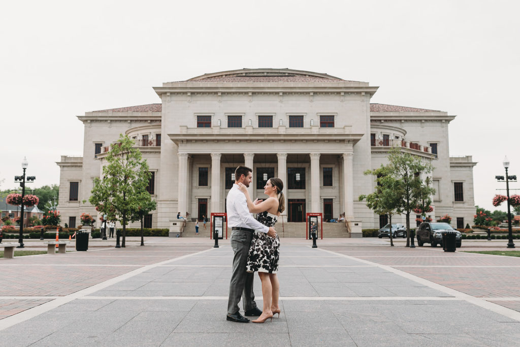 a man and woman hug and dance in front of the palladium in this ultra wide shot during their Carmel City Center engagement session