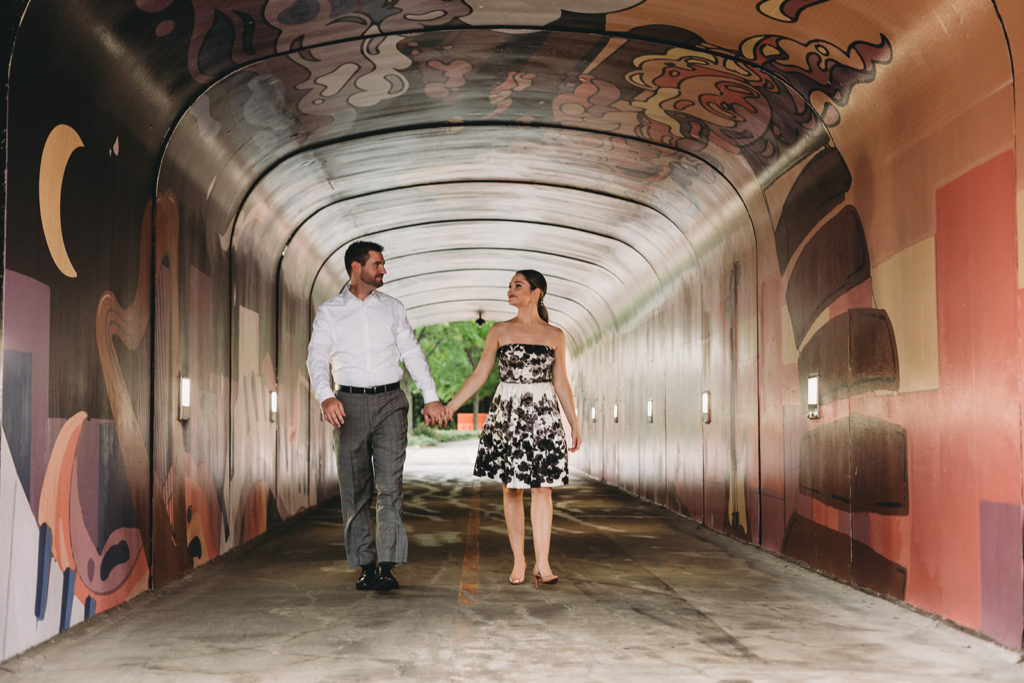 a man and a woman hold hands and walk through a tunnel covered in graffiti art during their Carmel City Center engagement session