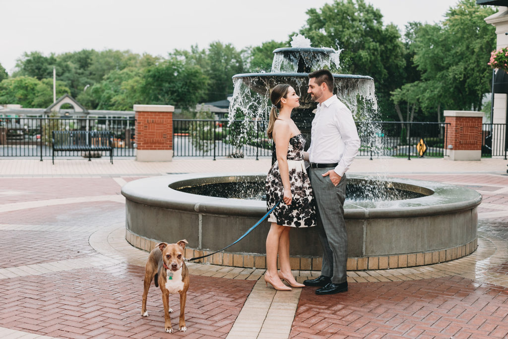 a man and woman in front of a fountain with their dog on a leash during their Carmel City Center engagement session