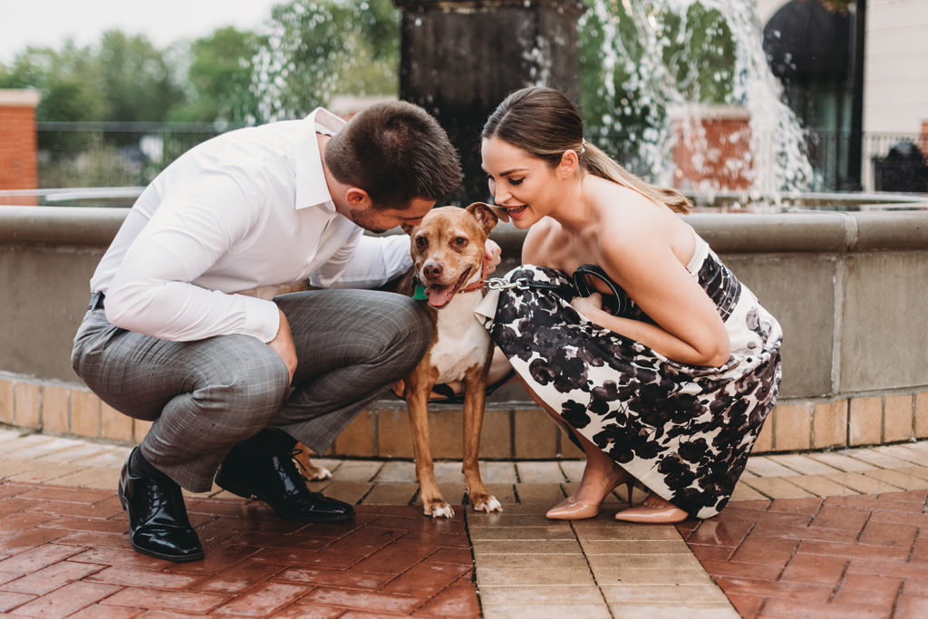 a man and woman kneel down to pet their dog in front of a fountain during their Carmel City Center engagement session