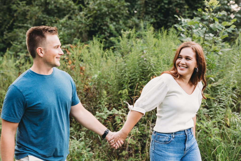 man and woman walk while holding hands in field during their eagle creek park engagement session