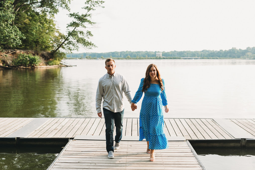woman in blue dress with white polka dots walks while holding hand with man on dock during their eagle creek park engagement session
