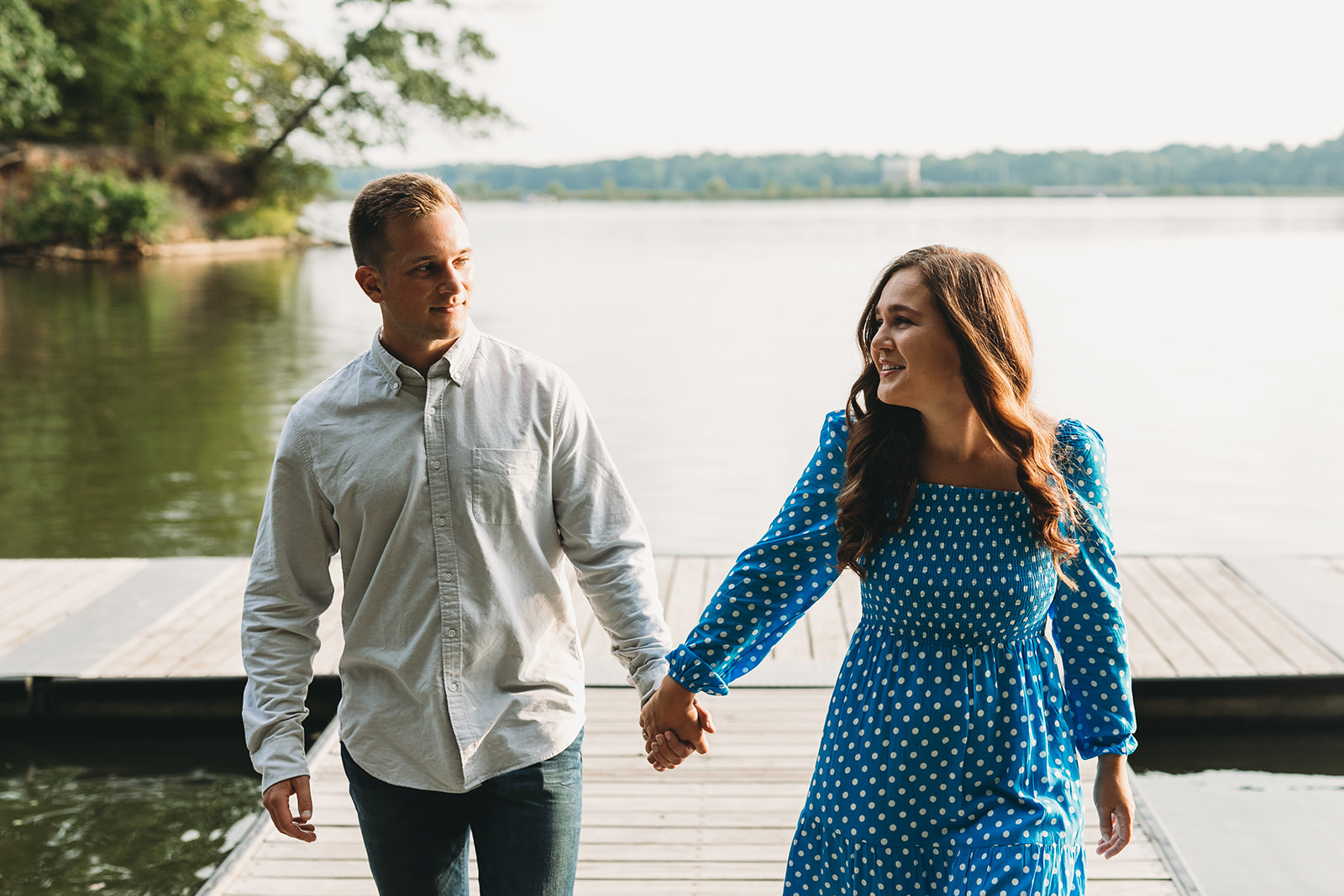 woman in blue dress and man in white collared shirt hold hands and walk done pier during their eagle creek park engagement session