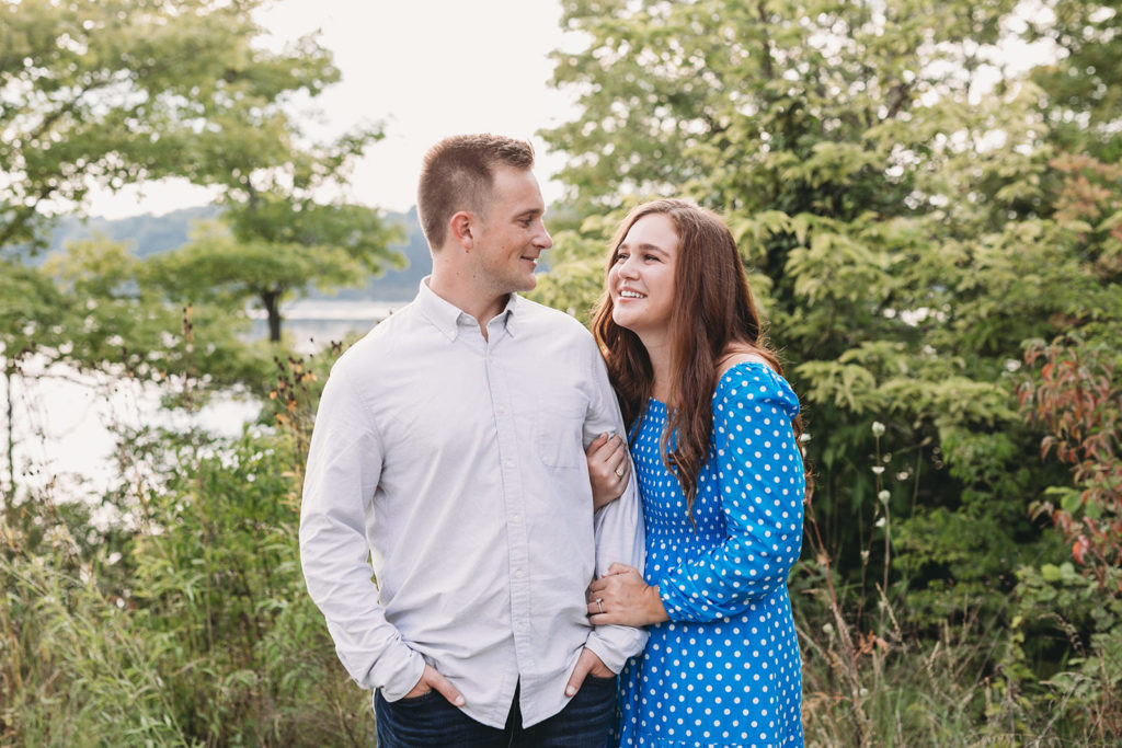 man with hands in pockets is arm hugged by woman in blue dress during their eagle creek park engagement session