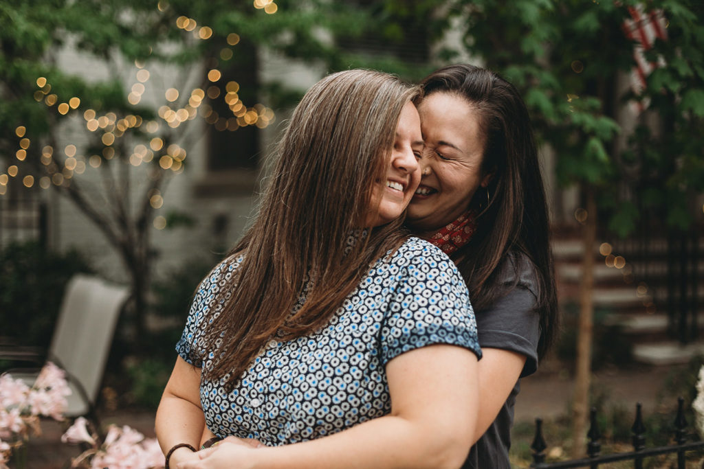 a woman hugs another woman from behind in front of bokeh during their lockerbie engagement session