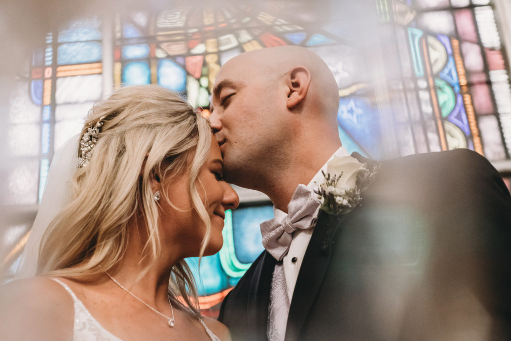 bride and groom kiss in front of stained glass during a marian university wedding