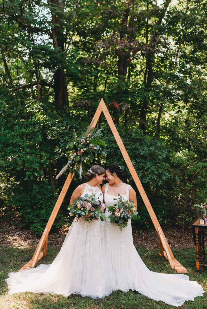 two brides touch foreheads in front of a wooden triangular arch during their Martinsville wedding