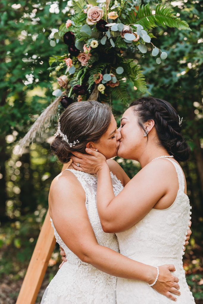 a bride with black hair grabs a bride with brown hair and kisses her on the lips while they hug during their Martinsville wedding