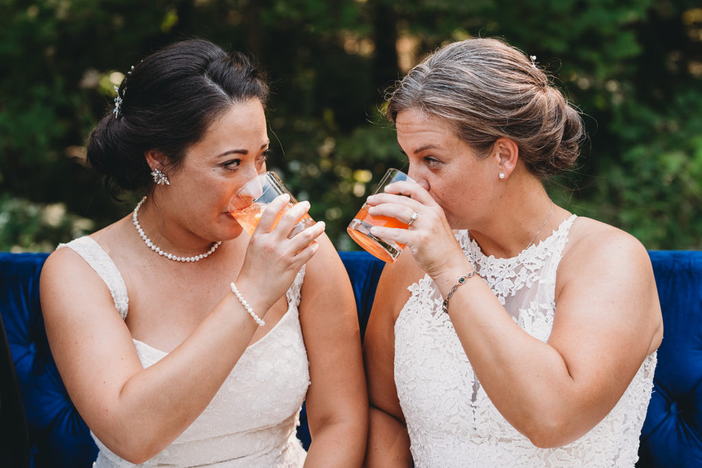 two brides sitting on an antique blue settee sipping signature cocktails while looking deep into each other's eyes during their Martinsville wedding