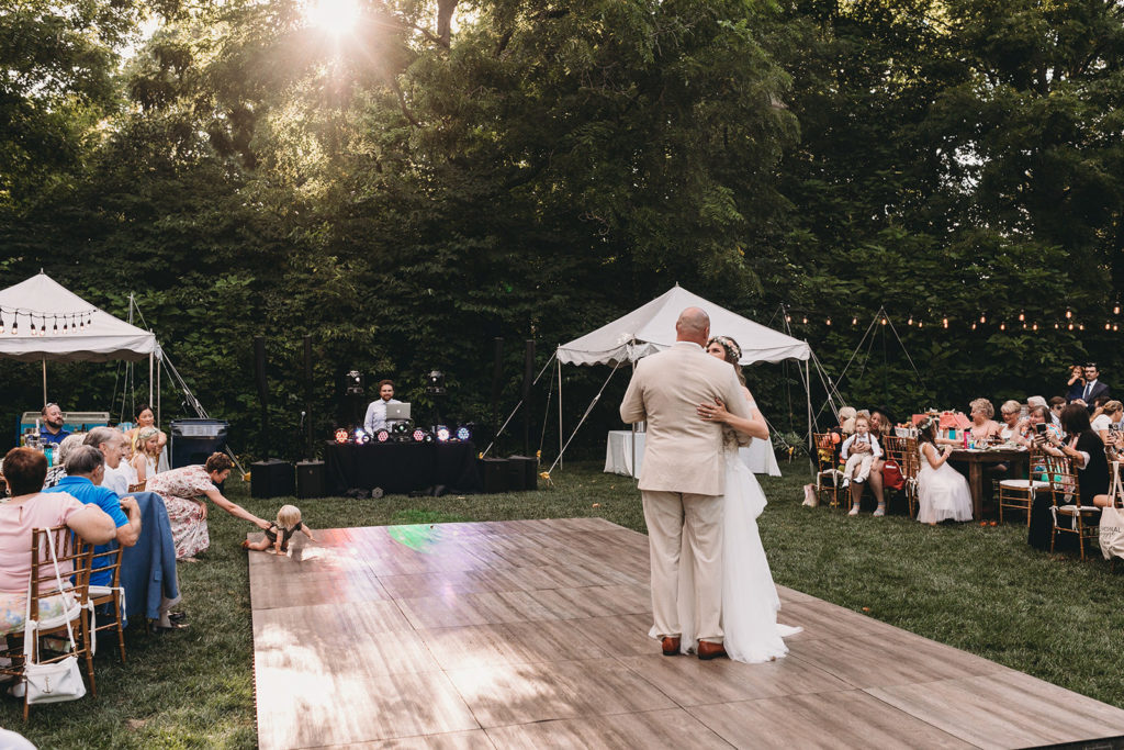 sun sets behind dancing bride and groom as a mom tries to keep her toddler from running out on the dance floor during the first dance during their Minnetrista wedding