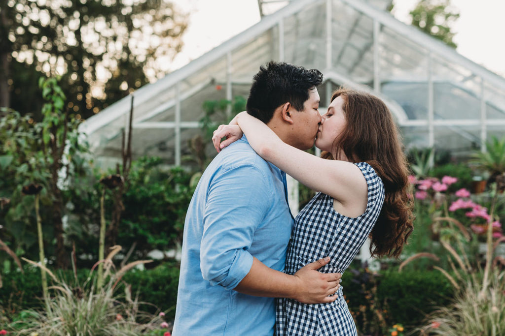 a man and a woman hug and kiss outside a glass building during their newfields greenhouse engagement photos