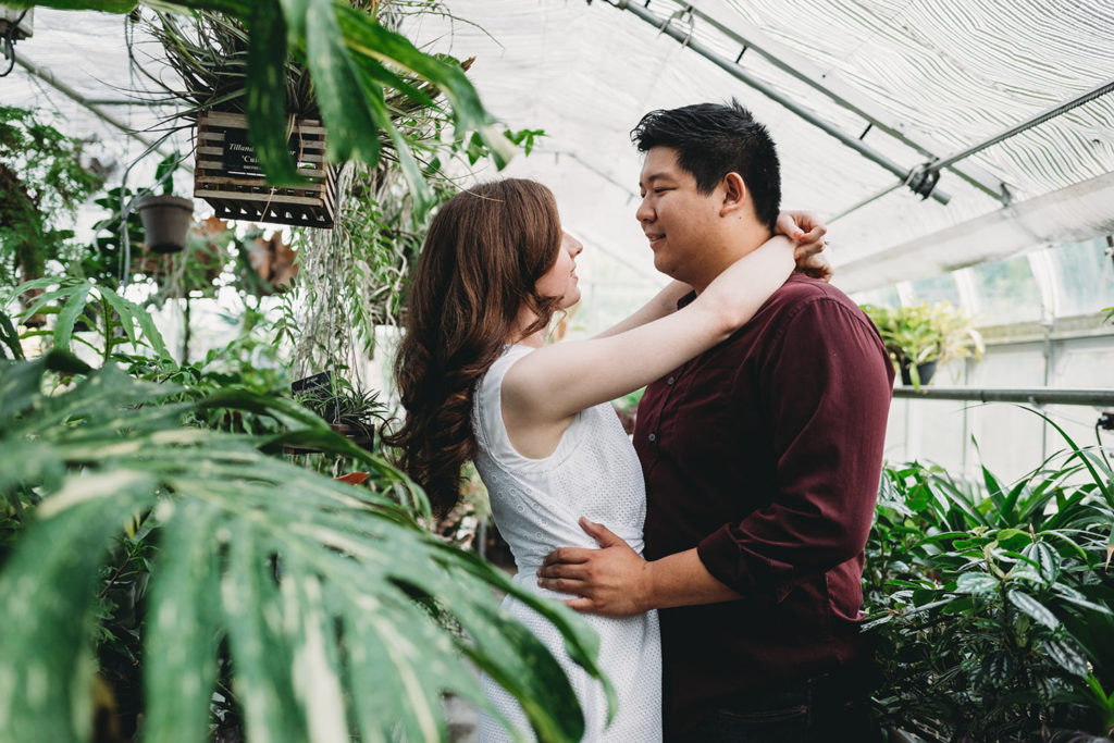 a woman hugs a man around the neck as he holds her waist in a glass building during their newfields greenhouse engagement photos