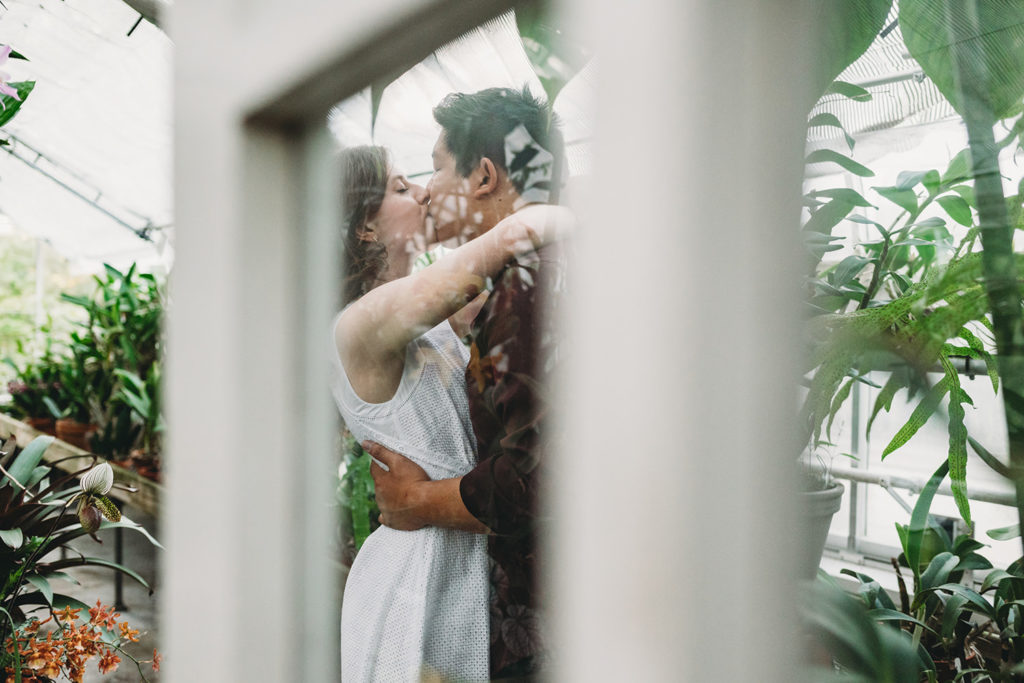 viewed through the glass of a door a man and a woman can be seen kissing inside a glass building during their newfields greenhouse engagement photos
