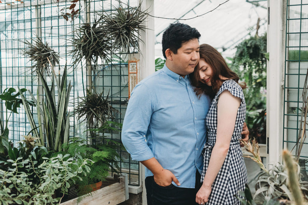 a man and a woman hug in front of plants during their newfields greenhouse engagement photos