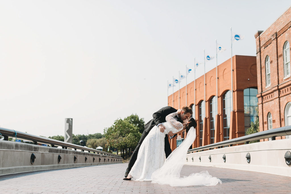 bride and groom kiss on a bridge in white river state park with the ncaa headquarters in the background before their scottish rite cathedral wedding