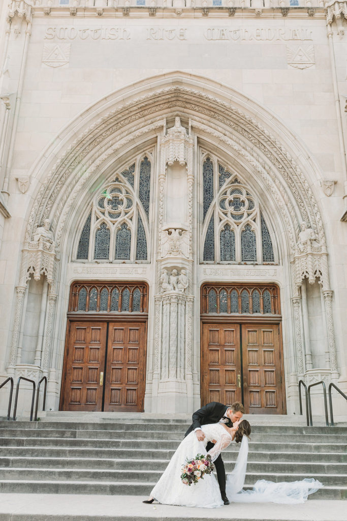 groom dips and kisses bride on steps outside the scottish rite cathedral main entrance during a scottish rite cathedral wedding