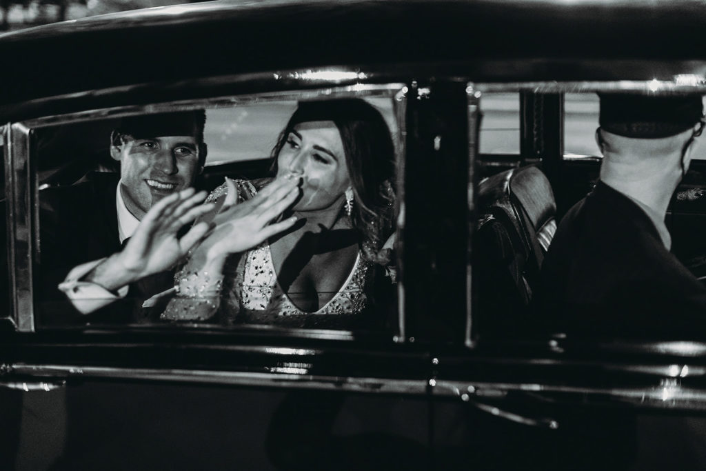 black and white photo of bride and groom looking out the window of a car and blowing kisses and waving as the car drives away at the end of their scottish rite cathedral wedding