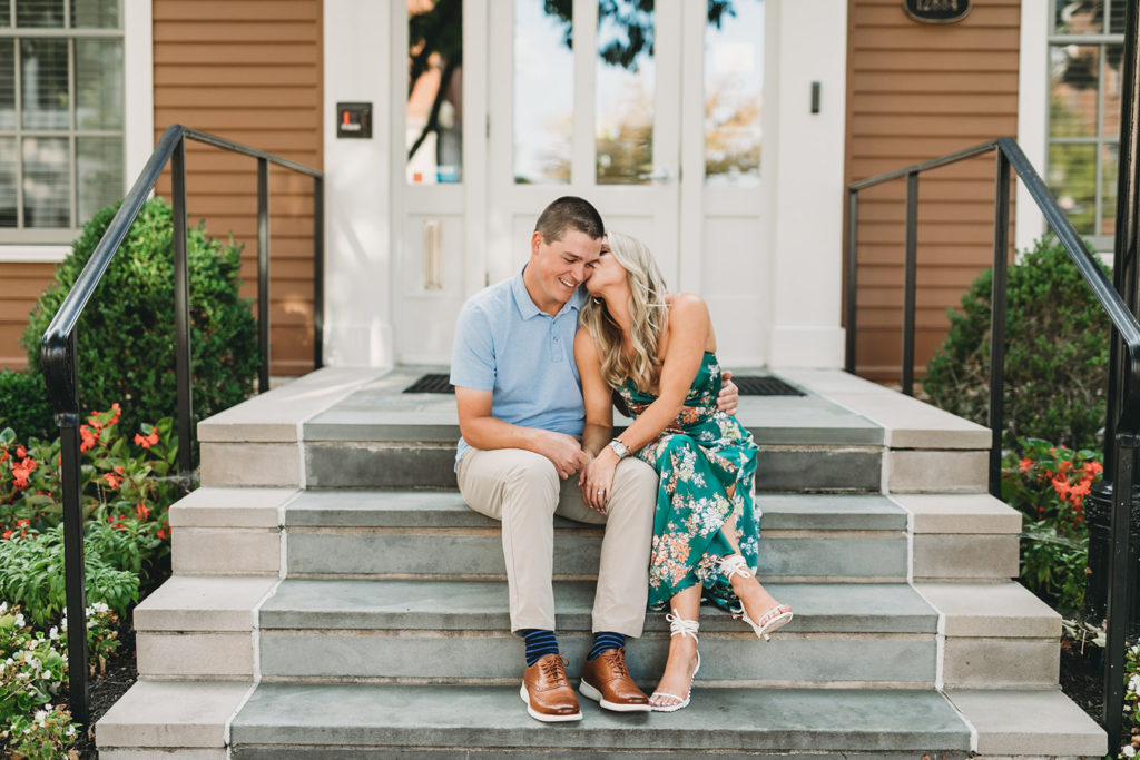 a woman in a green floral dress kisses a man in khakis and a polo shirt on the cheek while they sit on steps during their West Clay Engagement Session