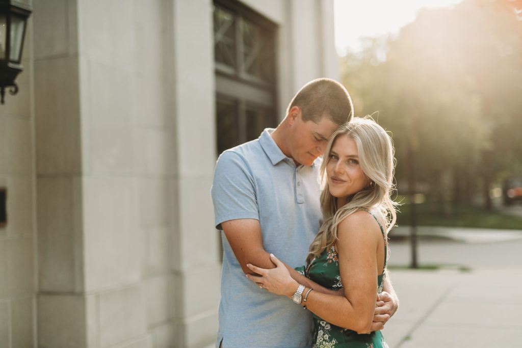 sun set behind woman in green floral dress with blond hair who is hugging. a man in a baby blue polo shirt during their West Clay Engagement Session 