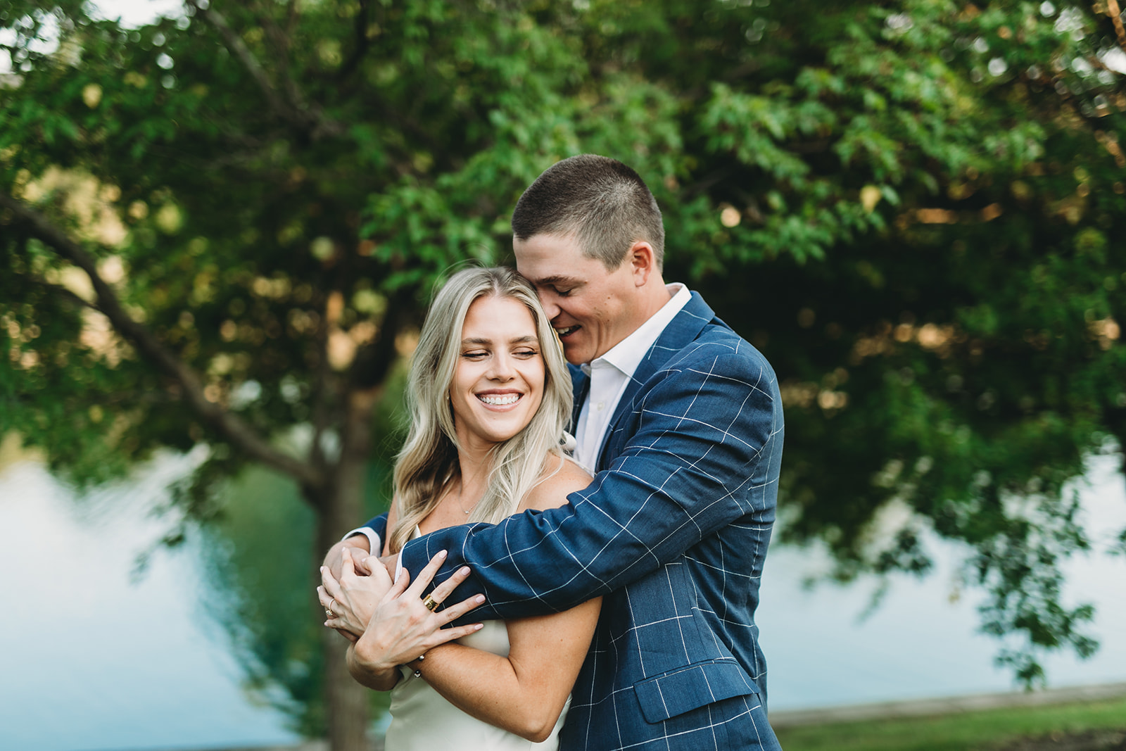 man in blue checked suit jacket hugs blonde woman while they both smile and laugh in front of a pond during their West Clay Engagement Session