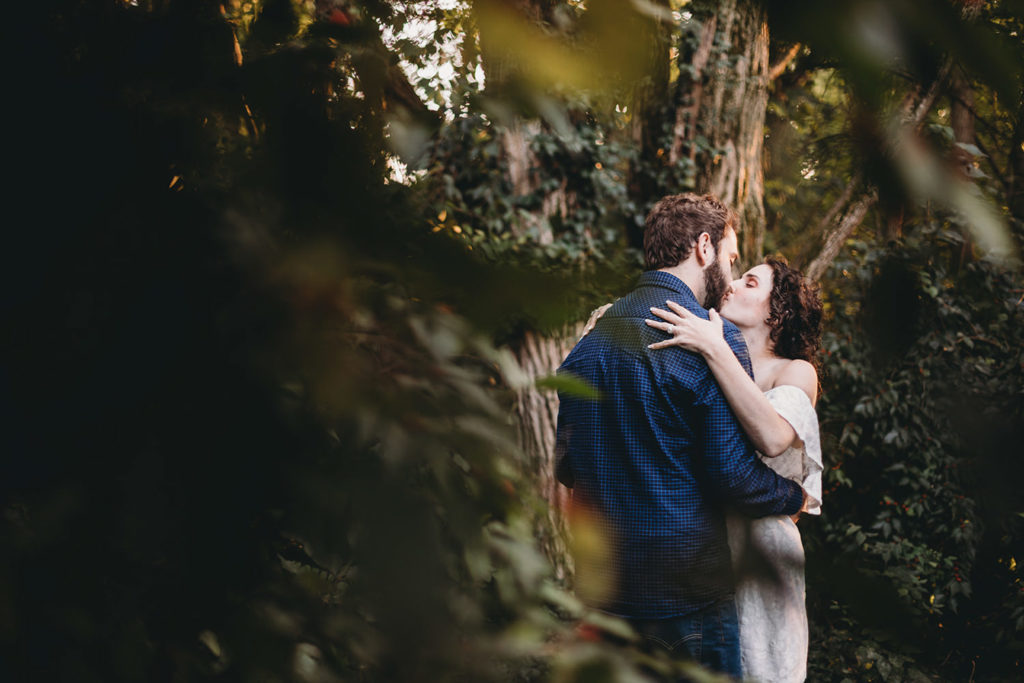viewed through a gap in the leaves of a bush, a man and a woman kiss during their Artisan Acres Estate engagement