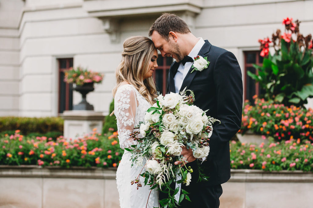 attractive bride and groom touch foreheads in front of flowers while hugging in carmel city center during their Artisan Acres Estate wedding