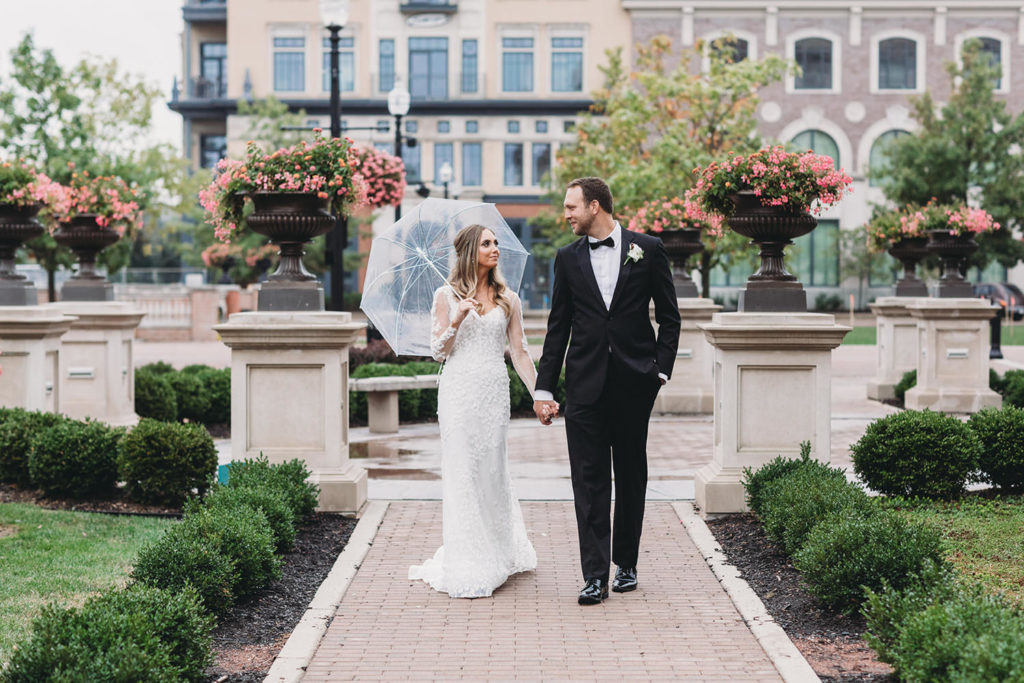 bride with clear umbrella talks walk with handsome groom in tux on rainy day in carmel city center during their Artisan Acres Estate wedding