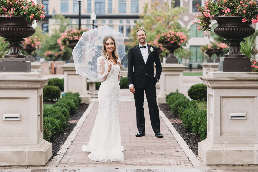 bride with umbrella stands on walkway while groom stands behind her smiling at carmel city center during their Artisan Acres Estate wedding