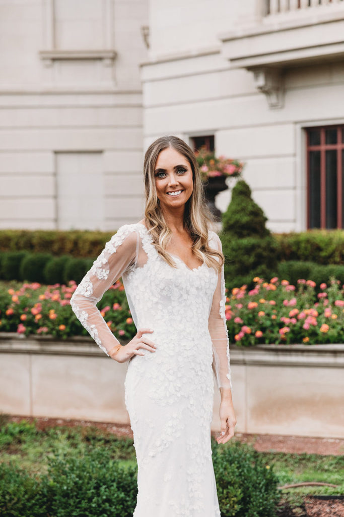 stunning bride in white dress with sheer sleeves stands in front of flowers in front of the palladium carmel cornerstone for the performing arts during their Artisan Acres Estate wedding