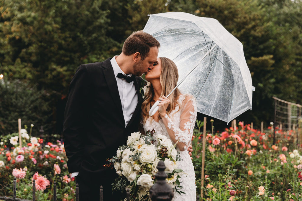 a man in a tux kisses a woman in a dress in front of flowers while she holds an umbrella to shelter them from the rain during their Artisan Acres Estate wedding