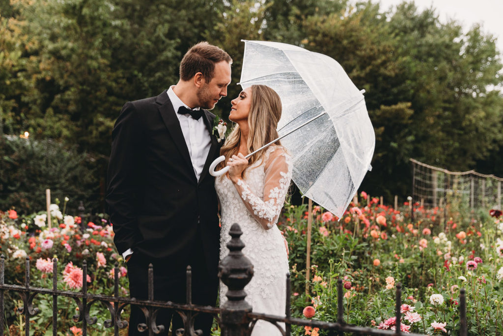 a man in a tux hugs a woman in a white dress holding a clear umbrella on a rainy day during their Artisan Acres Estate wedding