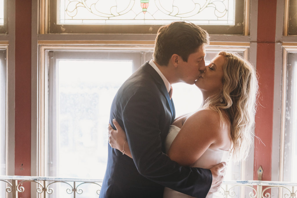 bride and groom backlit by stained glass window light kissing during their first look during their fountain square theatre wedding 