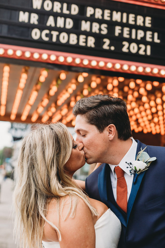 bride and groom kiss under the thousands of lights making up the theater marquee at sunset and the marquee says world premiere of the two of them during their fountain square theatre wedding
