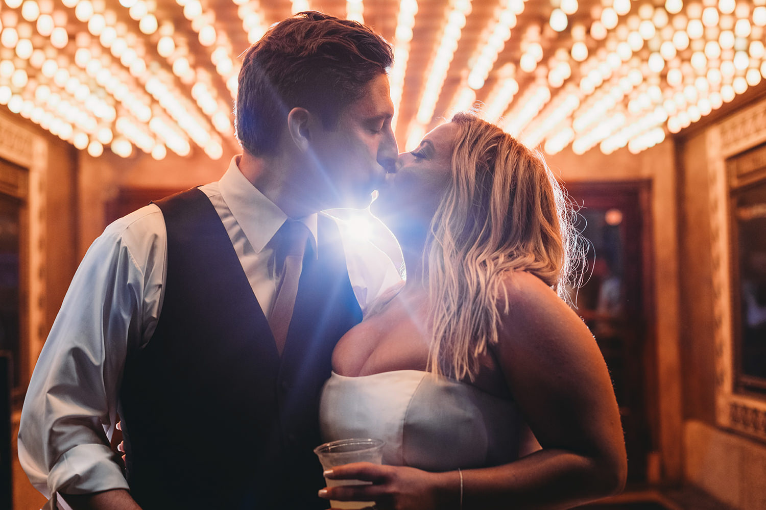 man and woman kiss while backlit by flash in front of lights of theater marquee during their fountain square theatre wedding