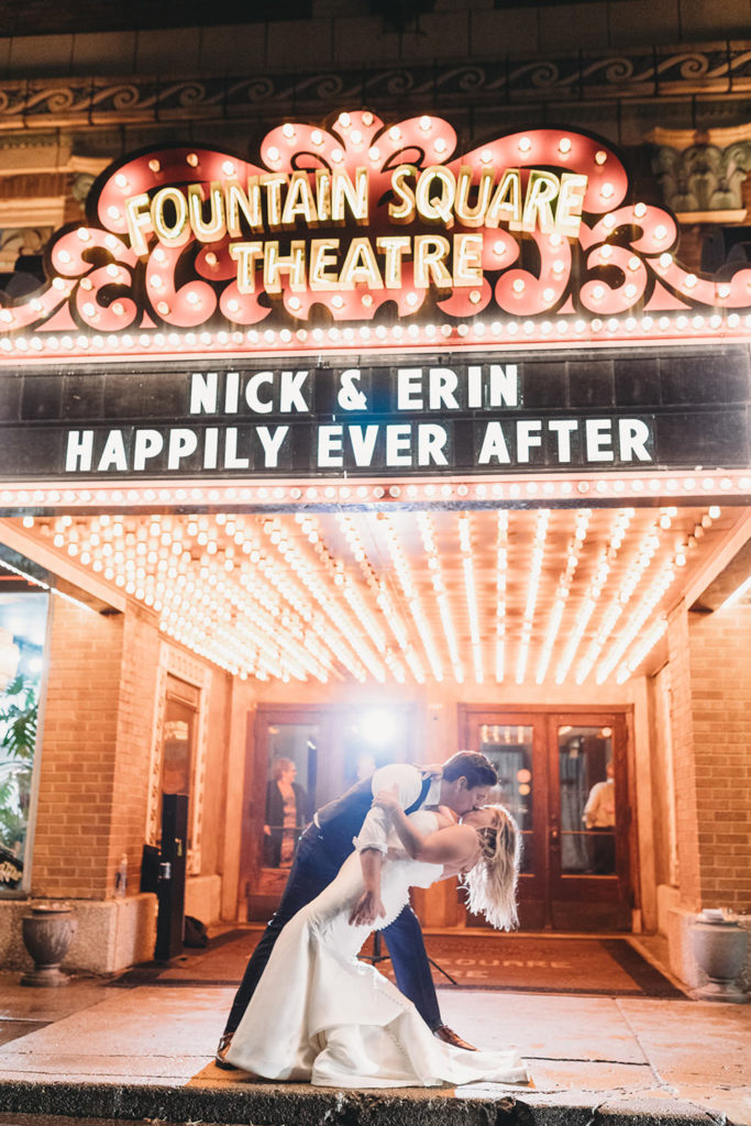 bride and groom do a dip and kiss under the theater marquee which says their names and happily ever after on it during their fountain square theatre wedding