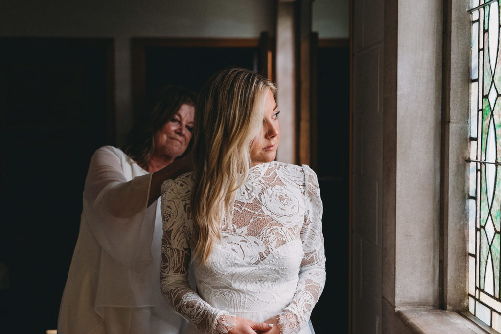 blonde bride looks out window as her mother buttons up her wedding dress at the graduate bloomington hotel during their indiana university wedding