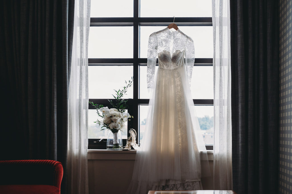a gorgeous wedding dress with a sheer lace top hangs in a window and backlit by the sun on a rainy day at the graduate bloomington hotel during their indiana university wedding