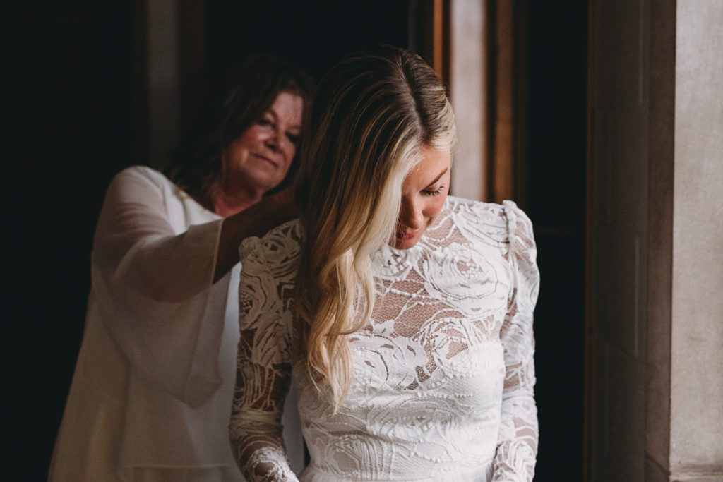 beautiful blonde bride is helped into a wedding dress by her mother at the graduate bloomington hotel during their indiana university wedding