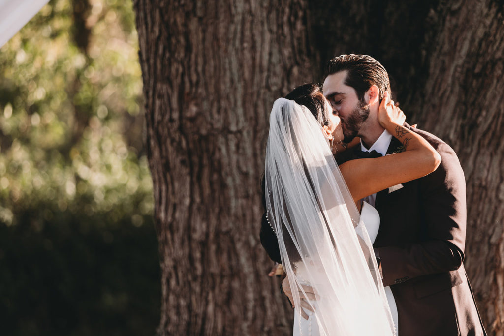 bride and groom first kiss in front of giant tree during their mustard seed wedding