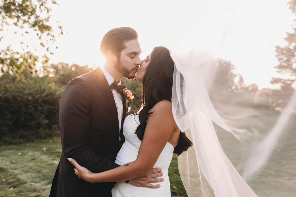 bride and groom kiss while veil blows in wind at sunset during their mustard seed wedding