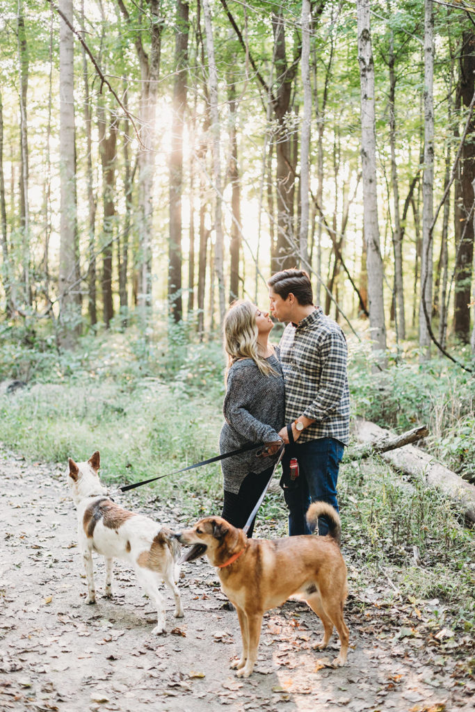 sun is setting behind trees as a man and a woman kiss while walking their dogs during their wonderful eagle creek engagement session
