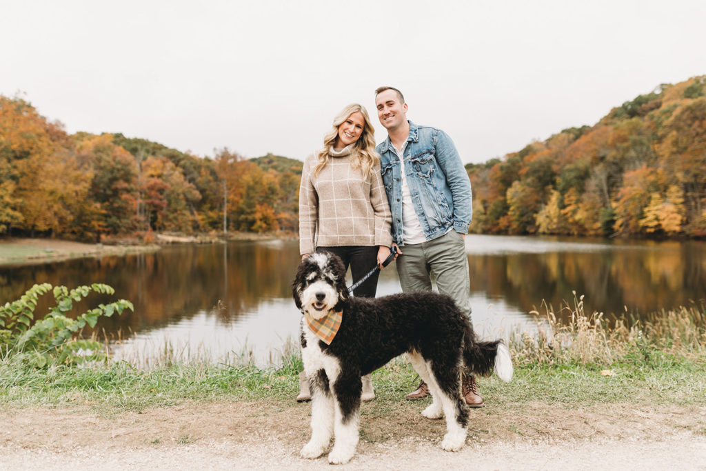 man and woman and big fluffy black and white dog in front of ogle lake in fall during their brown county engagement session